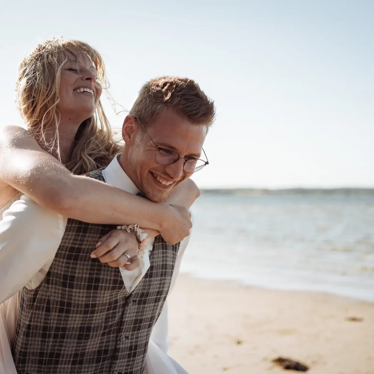 Junges Brautpaar lacht am Strand: Mann mit Brille trägt Frau huckepack, beide strahlen in der Sonne – echte Hochzeitsfotografie aus Rosenheim