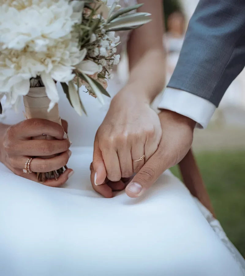 Nahaufnahme der Hände eines Brautpaares mit Trauringen, Braut hält weißen Brautstrauß mit grünen Blättern, natürliche und emotionale Hochzeitsfotografie